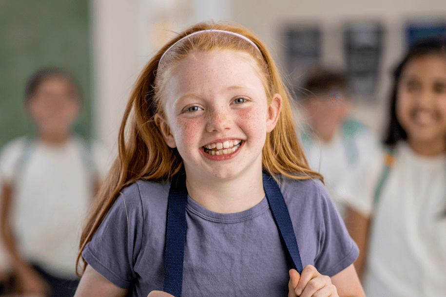 Une jeune fille qui souri. Elle va à son cours d'anglais à Genève. Elle a 8 ans, un t-shirt violet et des cheveux roux.