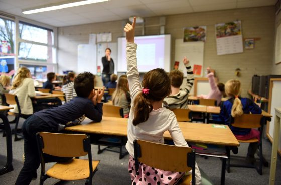 Une salle de classe remplie d'élève et d'un professeur. Une fille est assise et lève la main pour participer au cours.